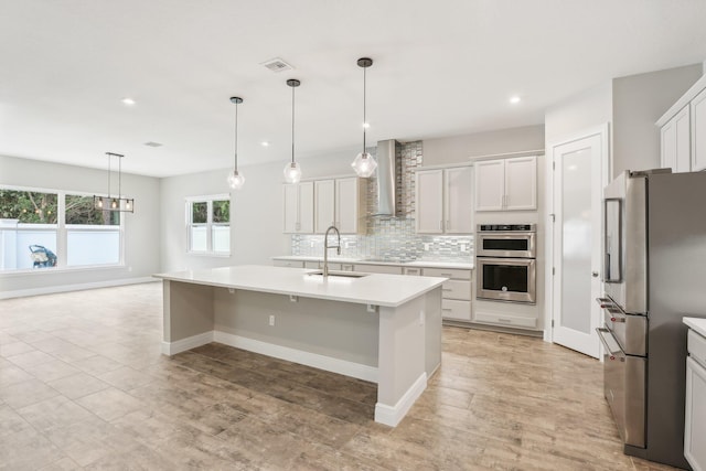 kitchen with sink, wall chimney exhaust hood, an island with sink, appliances with stainless steel finishes, and decorative light fixtures