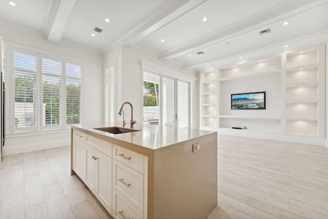 kitchen featuring sink, beam ceiling, light hardwood / wood-style floors, white cabinetry, and an island with sink