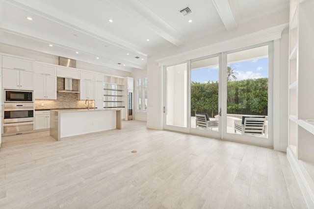 kitchen with black microwave, stainless steel oven, wall chimney range hood, beamed ceiling, and white cabinets