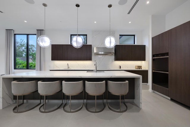 kitchen with backsplash, wall chimney exhaust hood, a kitchen island with sink, hanging light fixtures, and light tile patterned flooring