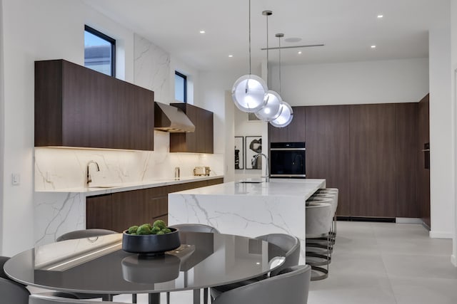 kitchen with a kitchen island with sink, oven, wall chimney range hood, sink, and decorative light fixtures