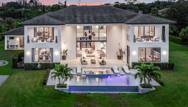 back house at dusk with a lawn, outdoor lounge area, a swimming pool with hot tub, a balcony, and a patio area