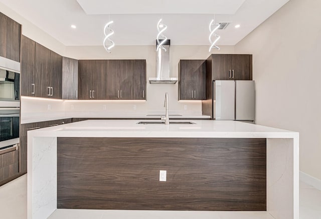 kitchen with a kitchen island with sink, white refrigerator, wall chimney exhaust hood, decorative light fixtures, and dark brown cabinetry