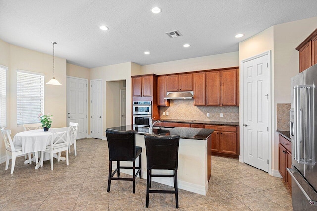 kitchen with dark countertops, under cabinet range hood, a center island with sink, and appliances with stainless steel finishes