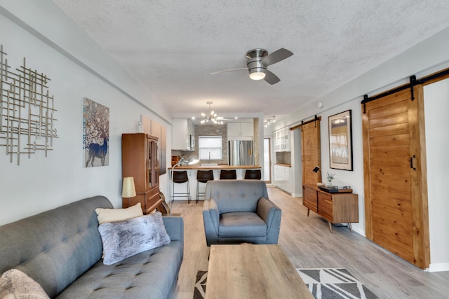 living room with ceiling fan with notable chandelier, a textured ceiling, a barn door, and light hardwood / wood-style flooring