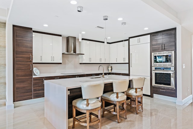 kitchen with wall chimney exhaust hood, dark brown cabinetry, sink, built in appliances, and hanging light fixtures