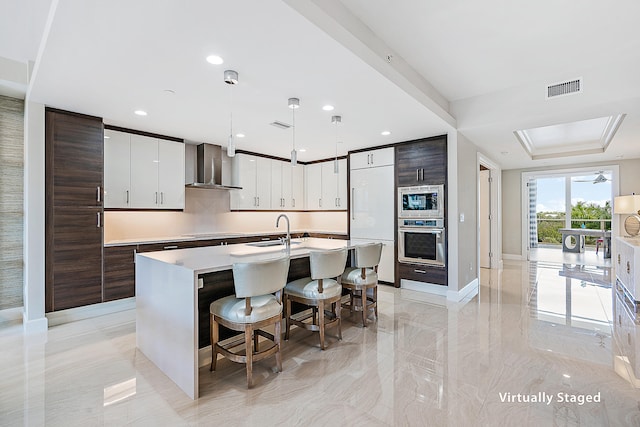 kitchen with wall chimney exhaust hood, a kitchen island with sink, built in appliances, hanging light fixtures, and a breakfast bar area