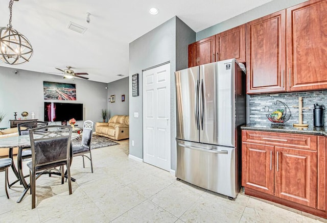 kitchen with backsplash, ceiling fan with notable chandelier, hanging light fixtures, dark stone countertops, and stainless steel refrigerator