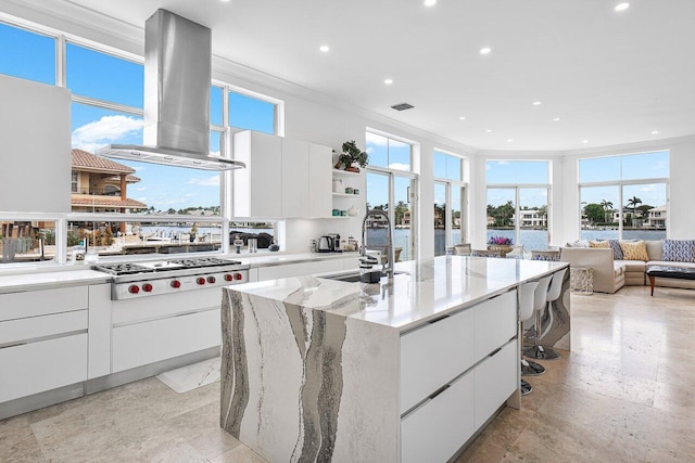 kitchen featuring stainless steel gas stovetop, a kitchen island with sink, wall chimney range hood, plenty of natural light, and white cabinetry