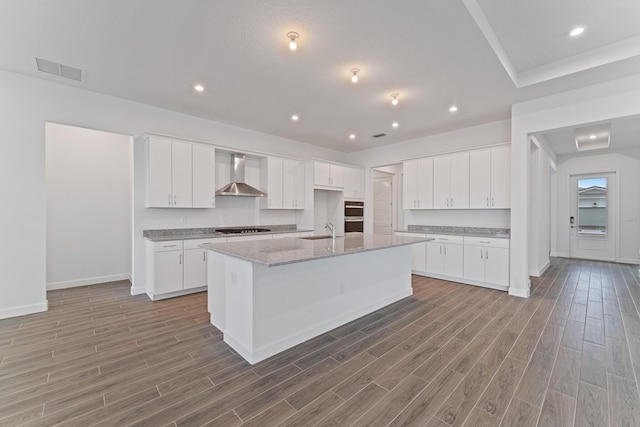 kitchen featuring wall chimney range hood, black gas stovetop, an island with sink, and white cabinets