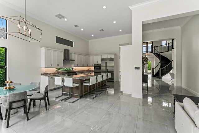 kitchen featuring wall chimney exhaust hood, crown molding, a kitchen bar, a center island with sink, and appliances with stainless steel finishes