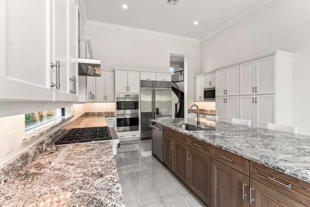 kitchen with light stone counters, ornamental molding, sink, built in appliances, and white cabinets