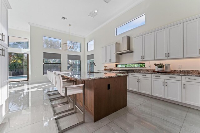 kitchen featuring a towering ceiling, wall chimney range hood, stone countertops, decorative light fixtures, and an island with sink