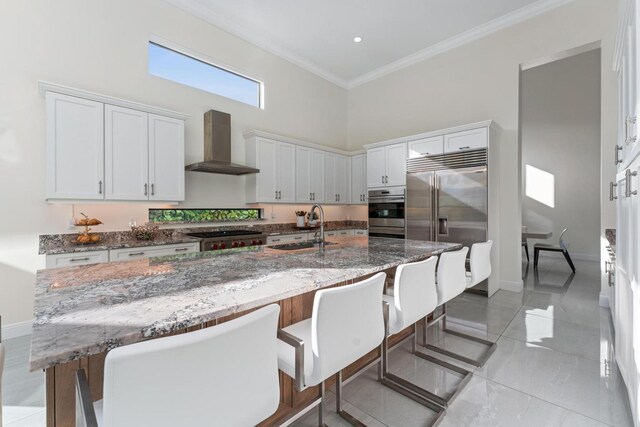 kitchen featuring a kitchen island with sink, a high ceiling, sink, wall chimney exhaust hood, and white cabinetry