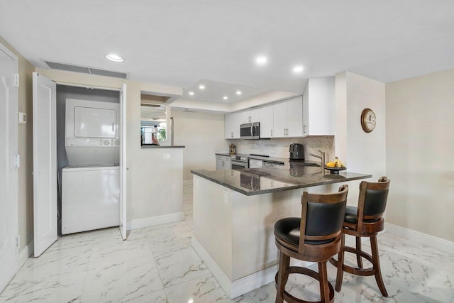 kitchen with kitchen peninsula, backsplash, stainless steel appliances, stacked washer and clothes dryer, and white cabinets