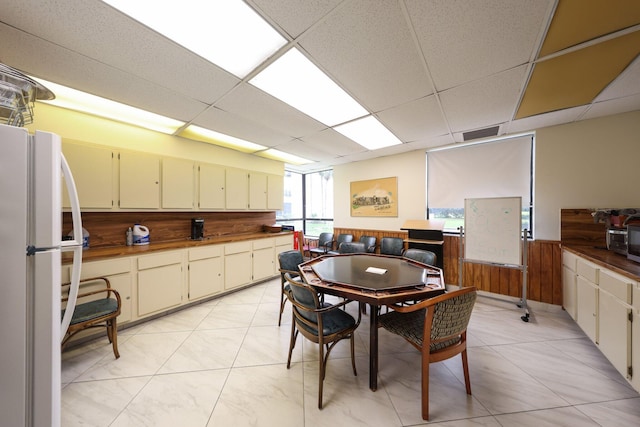 kitchen with cream cabinets, white refrigerator, a drop ceiling, and wood walls