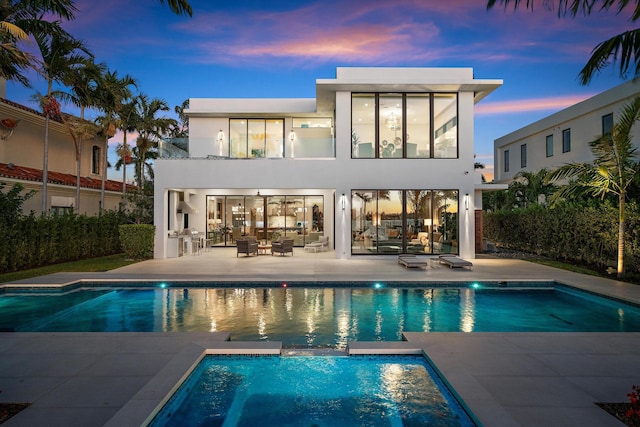 back house at dusk featuring a pool with hot tub, a patio area, and a balcony