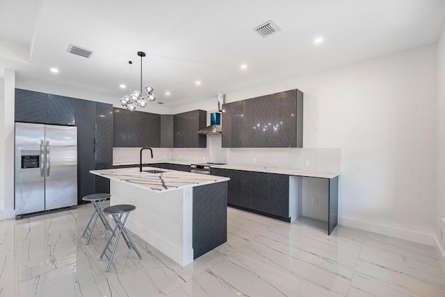 kitchen with appliances with stainless steel finishes, wall chimney exhaust hood, a kitchen island with sink, sink, and decorative light fixtures