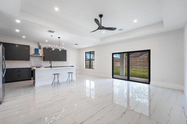 interior space featuring ceiling fan, wall chimney exhaust hood, a tray ceiling, a center island with sink, and a kitchen bar