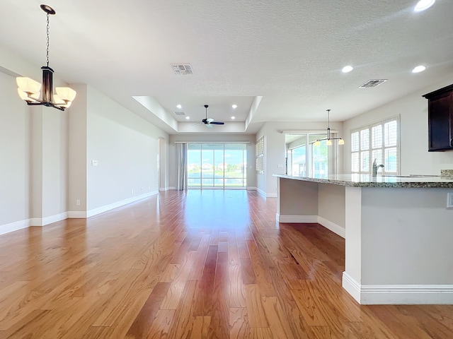 unfurnished living room with a raised ceiling, ceiling fan with notable chandelier, light hardwood / wood-style flooring, and a healthy amount of sunlight