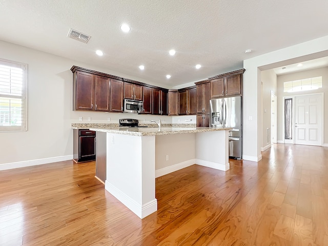 kitchen featuring light stone counters, a textured ceiling, stainless steel appliances, light hardwood / wood-style flooring, and an island with sink