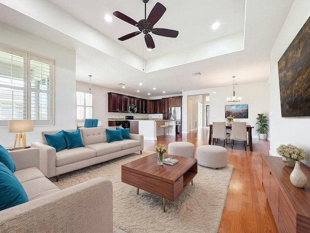 living room featuring a tray ceiling, ceiling fan with notable chandelier, and light wood-type flooring