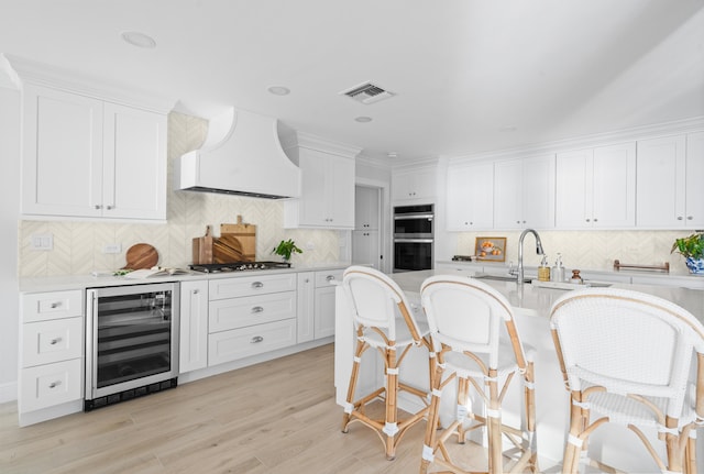 kitchen with white cabinetry, custom exhaust hood, stainless steel appliances, light wood-type flooring, and beverage cooler