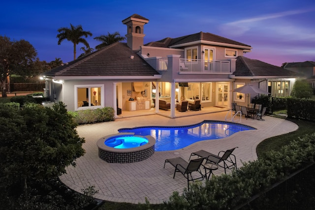 back house at dusk featuring a balcony, an outdoor kitchen, an outdoor hangout area, a pool with hot tub, and a patio area