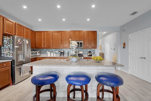 kitchen with appliances with stainless steel finishes, light wood-type flooring, tasteful backsplash, a breakfast bar, and a kitchen island with sink