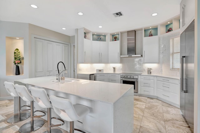 kitchen with backsplash, a kitchen island with sink, wall chimney range hood, sink, and appliances with stainless steel finishes