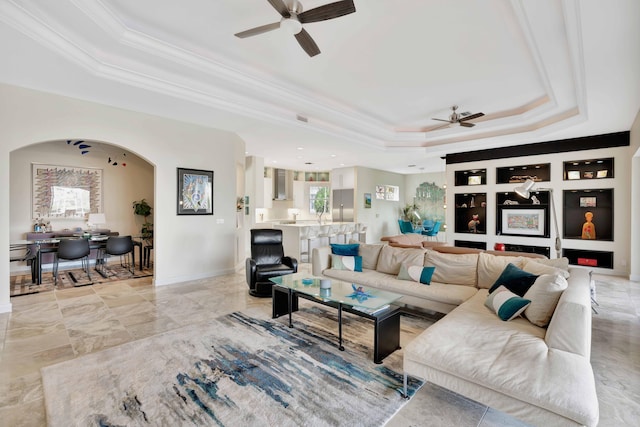 living room with a raised ceiling, ceiling fan, built in features, and ornamental molding