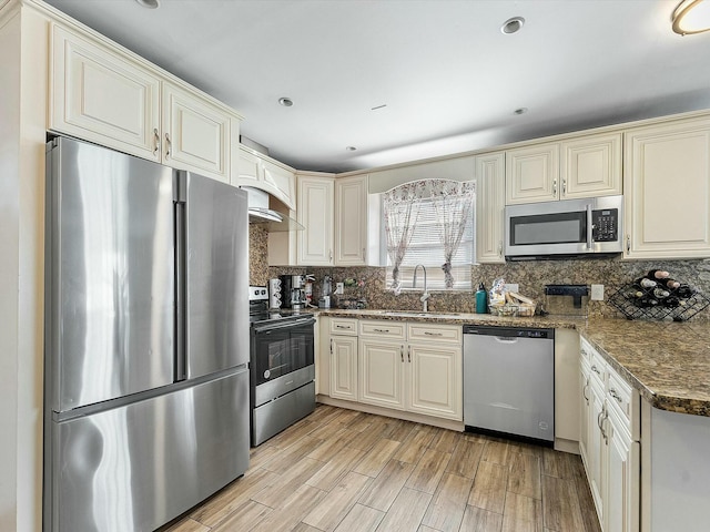 kitchen featuring appliances with stainless steel finishes, light wood-type flooring, dark stone counters, exhaust hood, and sink