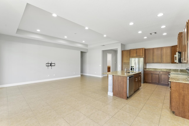 kitchen featuring a tray ceiling, light stone counters, a center island with sink, and appliances with stainless steel finishes