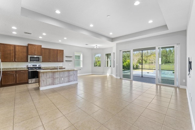 kitchen featuring stainless steel appliances, a raised ceiling, light stone counters, a kitchen island with sink, and light tile patterned floors