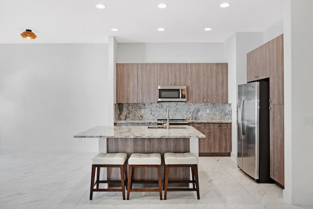 kitchen featuring light stone countertops, decorative backsplash, a kitchen bar, stainless steel appliances, and an island with sink