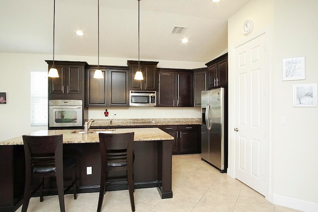 kitchen with light stone countertops, stainless steel appliances, sink, decorative light fixtures, and an island with sink