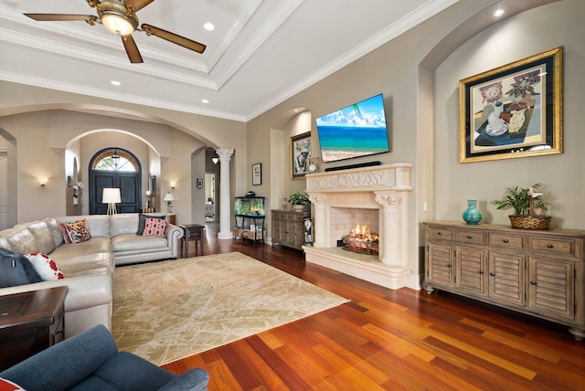 living room with a high end fireplace, ornamental molding, a tray ceiling, ceiling fan, and dark wood-type flooring