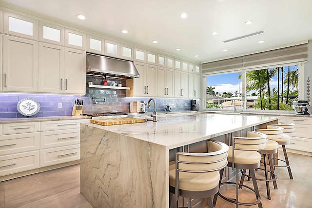 kitchen featuring light stone counters, white cabinetry, an island with sink, and exhaust hood