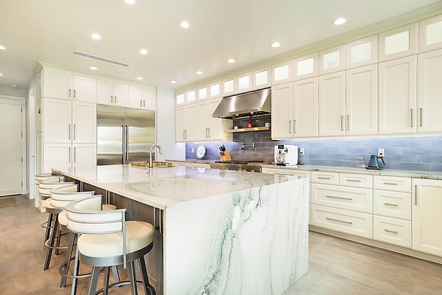 kitchen featuring white cabinetry, light stone counters, range hood, a large island with sink, and appliances with stainless steel finishes