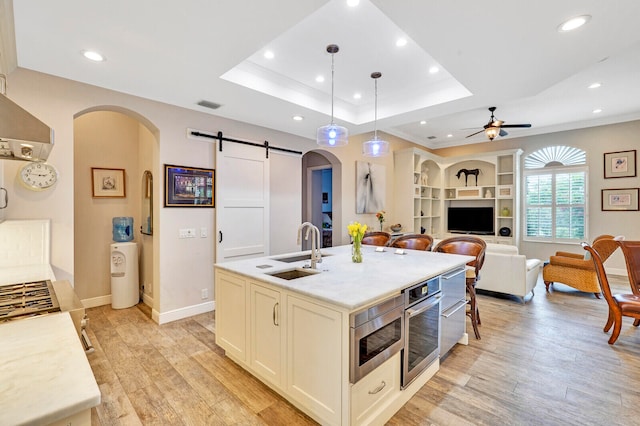 kitchen with light wood-type flooring, a kitchen island with sink, sink, a barn door, and hanging light fixtures