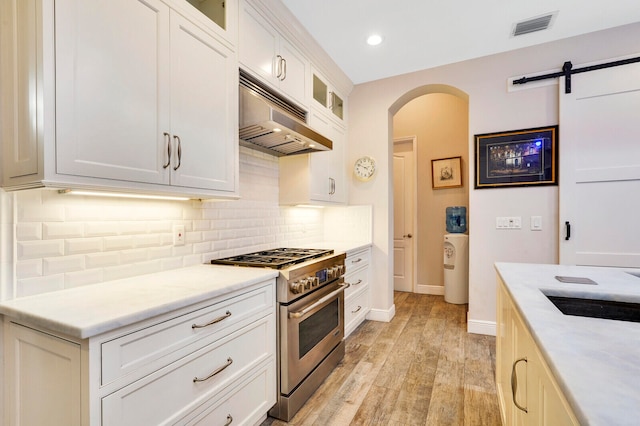 kitchen with stainless steel stove, tasteful backsplash, a barn door, light hardwood / wood-style flooring, and white cabinets