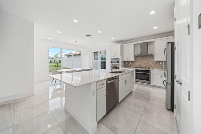 kitchen featuring hanging light fixtures, wall chimney exhaust hood, an island with sink, white cabinetry, and stainless steel appliances