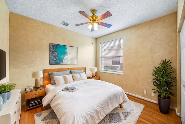 bedroom with a textured ceiling, light wood-style flooring, a ceiling fan, visible vents, and baseboards