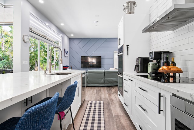 kitchen featuring white cabinetry, wall chimney exhaust hood, stainless steel appliances, light stone counters, and light hardwood / wood-style floors