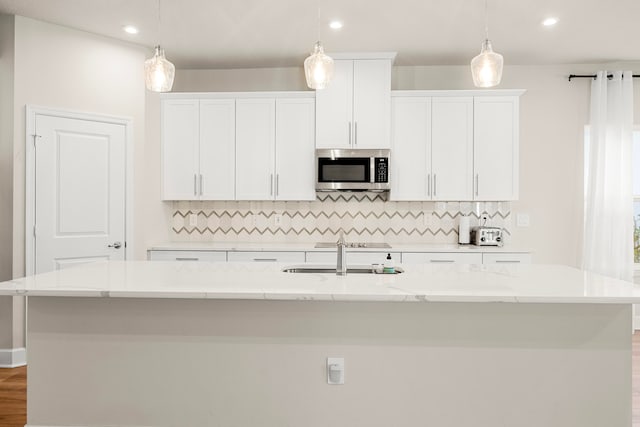 kitchen with tasteful backsplash, light stone counters, pendant lighting, white cabinets, and an island with sink