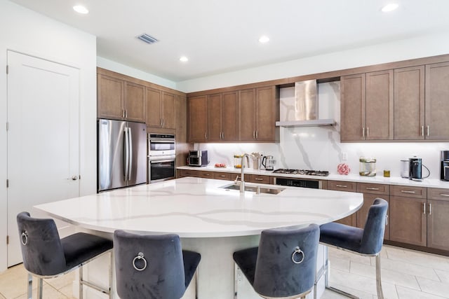 kitchen featuring wall chimney range hood, appliances with stainless steel finishes, a kitchen bar, and an island with sink