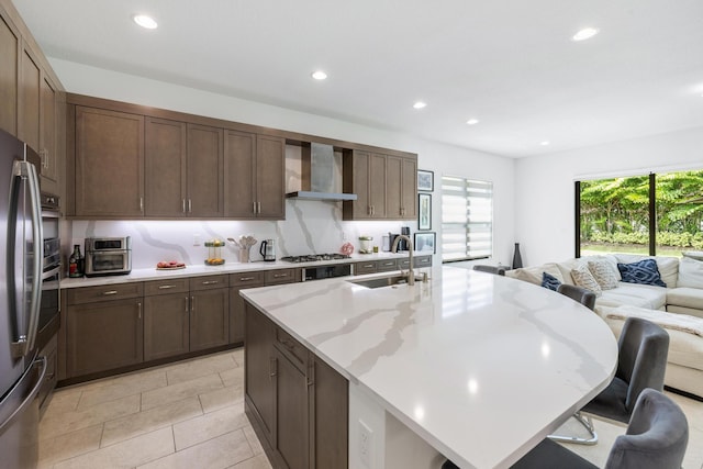 kitchen with appliances with stainless steel finishes, light stone counters, wall chimney exhaust hood, sink, and a center island with sink