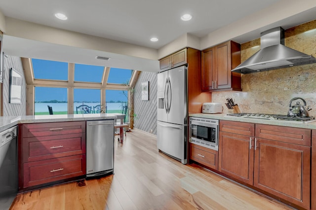 kitchen featuring backsplash, stainless steel appliances, wall chimney range hood, a water view, and light hardwood / wood-style floors