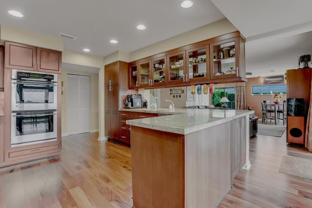 kitchen with kitchen peninsula, light wood-type flooring, stainless steel double oven, and light stone countertops