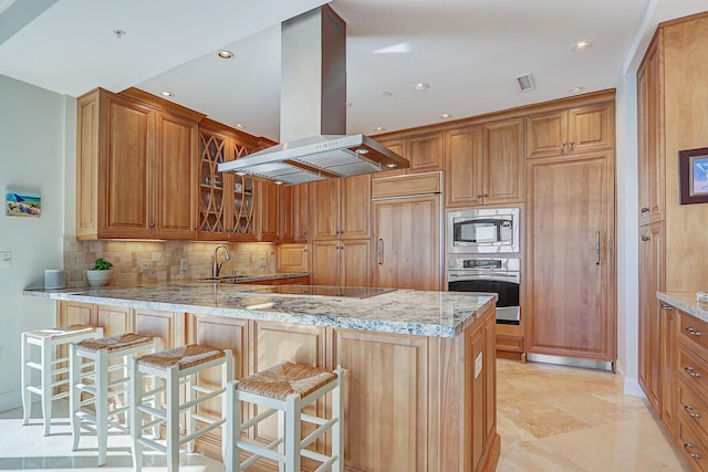 kitchen with built in appliances, kitchen peninsula, light stone counters, island exhaust hood, and a kitchen breakfast bar